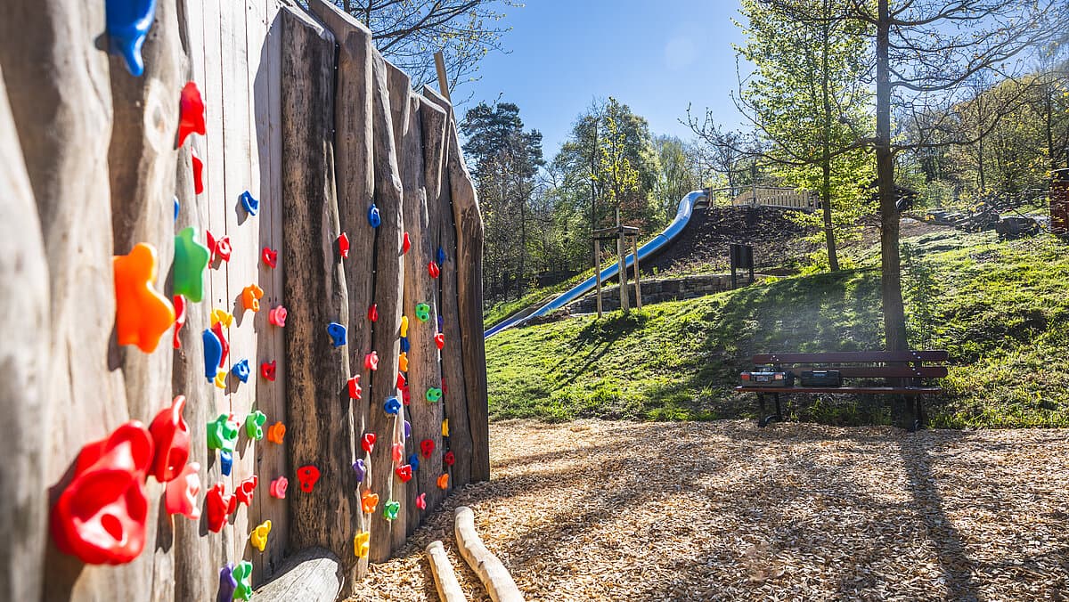 Links eine Kletterwand für Kinder mit bunten Haltesteinen, im Hintergrund eine Rutsche inmitten von Bäumen. © Stadt Gera