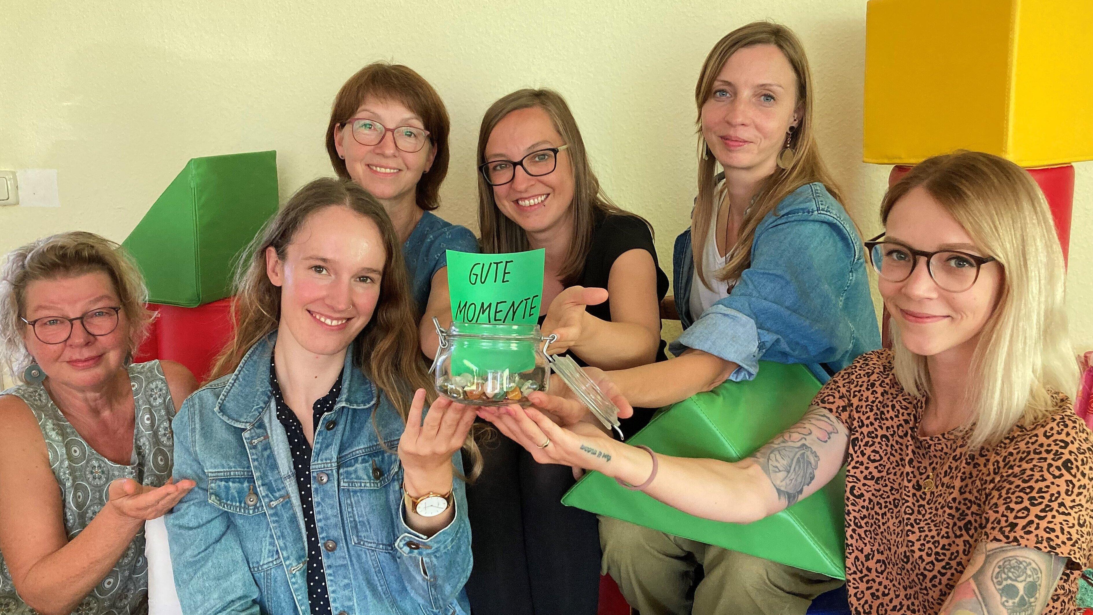 Foto zeigt das Team der Erziehungsberatung der Trägerwerke Soziale Dienste in Gera. Sie halten ein Glas in der Hand mit dem Zettel und der Aufschrift "Gute Momente". Sie sitzen als Gruppe zwischen bunten geometrischen Figuren..